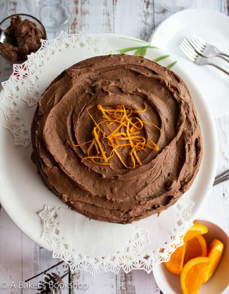 Overhead shot of a round chocolate orange cake sitting on a white cake stand. A small bowl of sliced oranges sits off to the side.