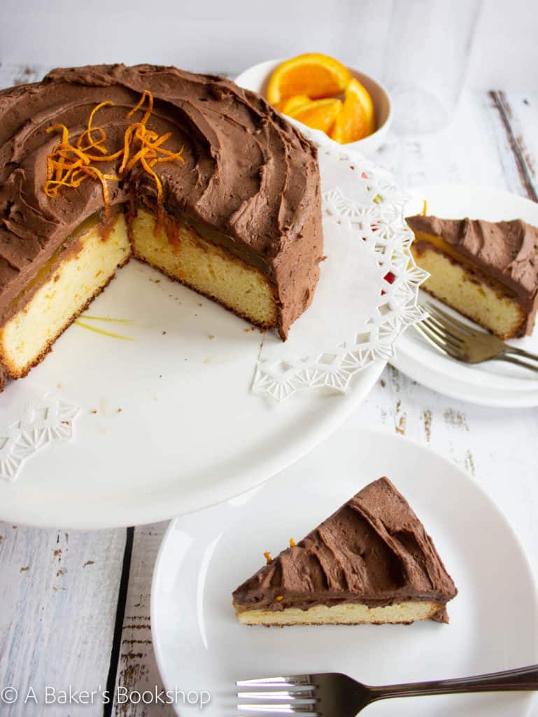 orange chocolate cake sits on a white cake stand with two slices of cake sitting to the side on white plates.  A small bowl of sliced oranges sits to the back.
