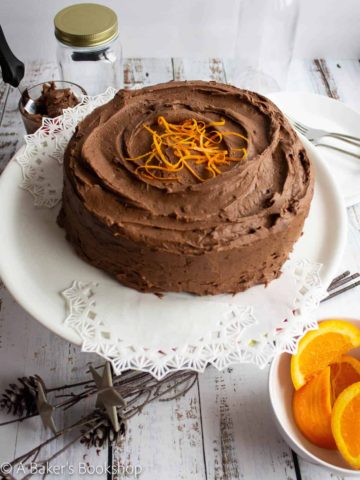 chocolate orange cake on white cake stand with a bowl of sliced oranges on the whiteside